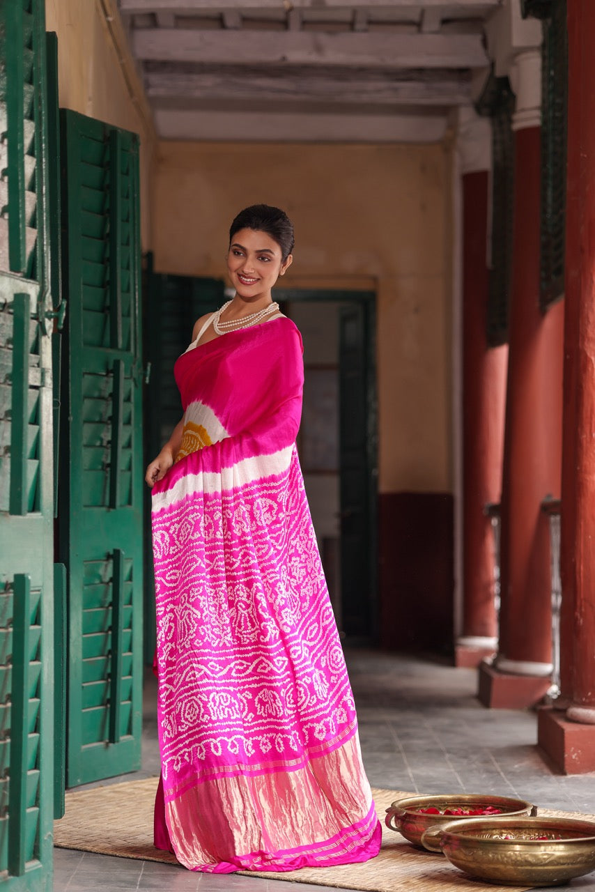 Woman in a pink saree with white patterns standing in a traditional setting.