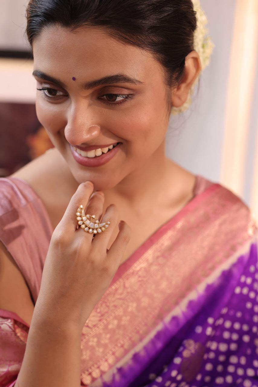 Woman wearing a purple saree with a gold ring on her finger, smiling.