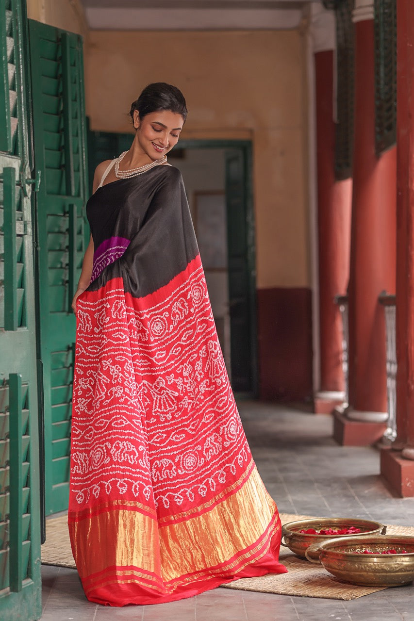 Woman in a black and red saree standing in a traditional setting with green doors and red walls.