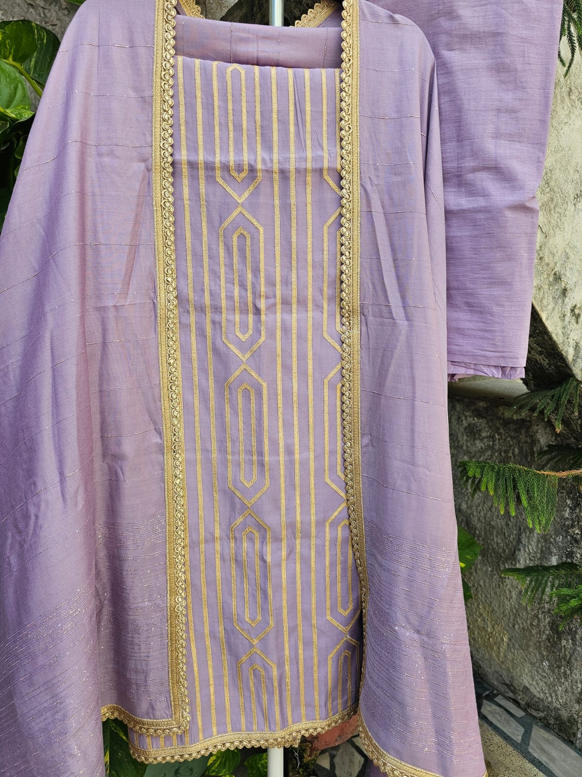 A lavender three-piece unstitched suit set with machine applique, consisting of a top, bottom, and dupatta, displayed against a natural background.