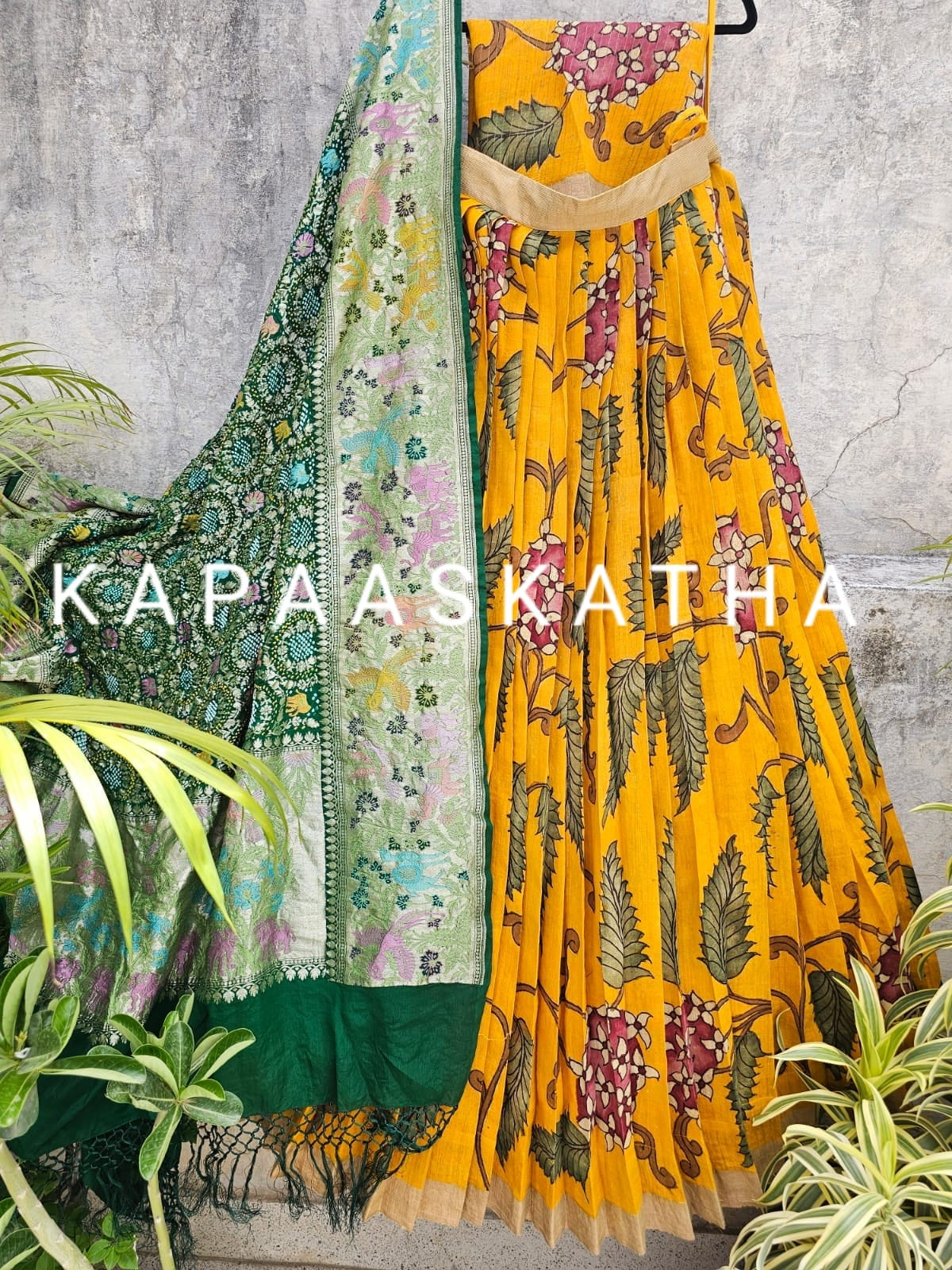 Yellow floral dress with green dupatta on a textured wall background, featuring the brand 'Kapaaskatha'.