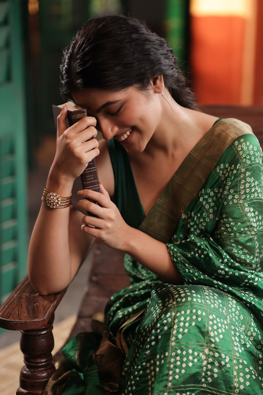 Woman in a green saree holding a cup, sitting on a wooden bench wearing a 92.5 silver handcuff with moissanite polki.