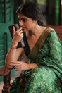 Woman in a green saree holding a cup, sitting on a wooden bench wearing a 92.5 silver handcuff with moissanite polki.