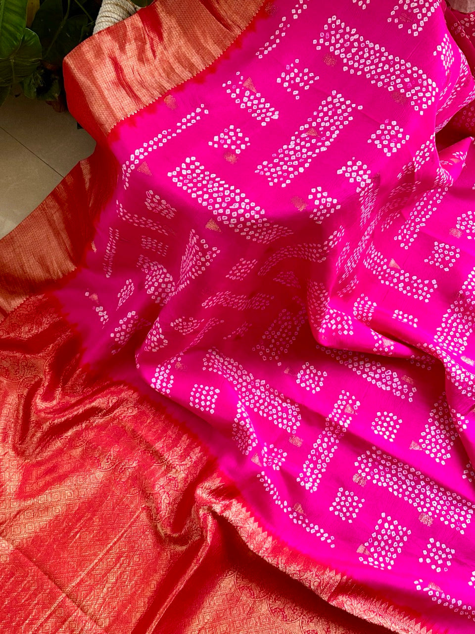 A rolled-up image of a red and pink Kanchipattu Bandhani saree with a geometric grid pattern and zari border detailing.