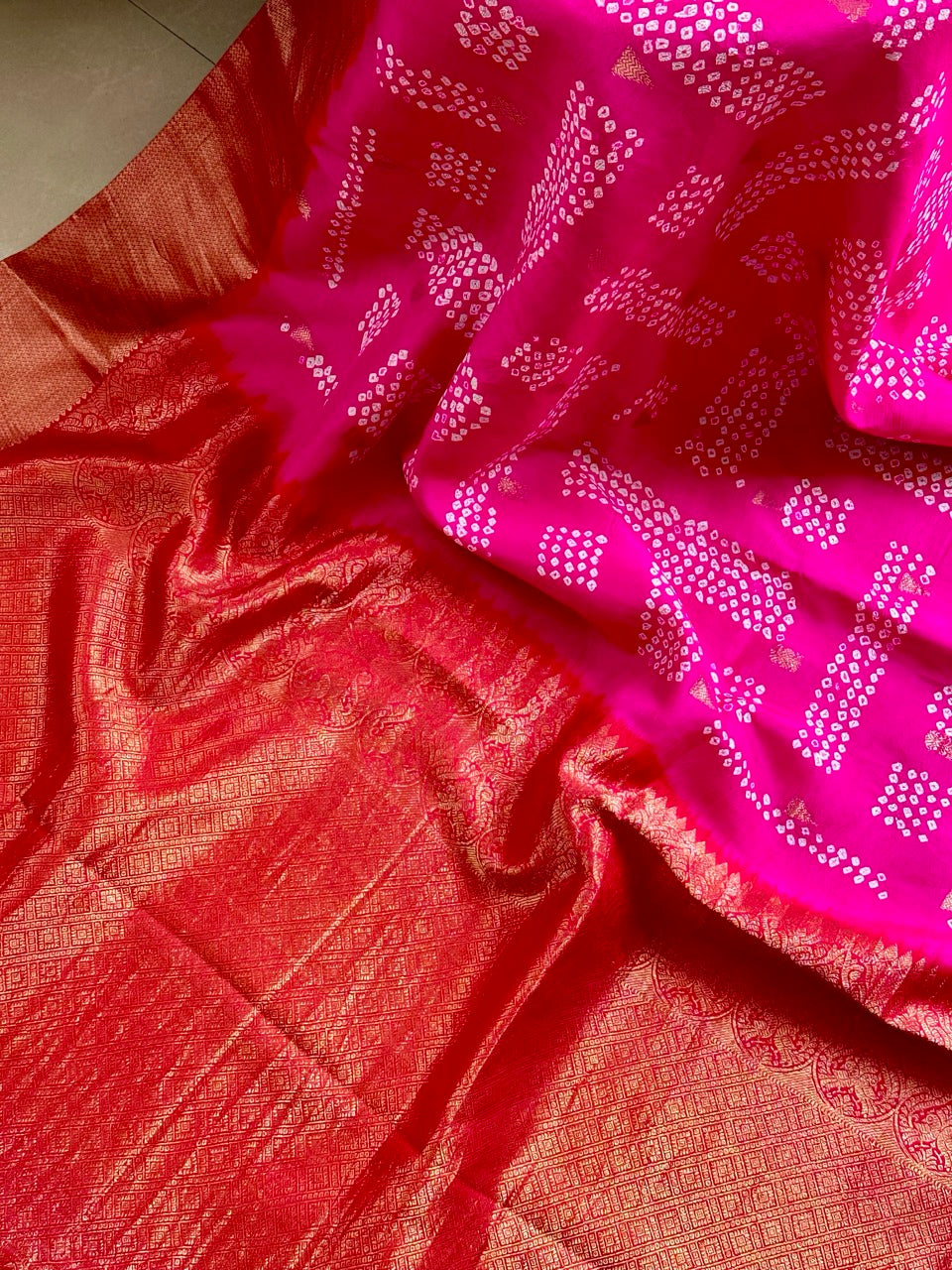 A rolled-up image of a red and pink Kanchipattu Bandhani saree with a geometric grid pattern and zari border detailing.