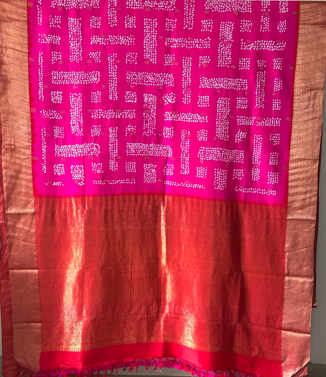 A rolled-up image of a red and pink Kanchipattu Bandhani saree with a geometric grid pattern and zari border detailing.