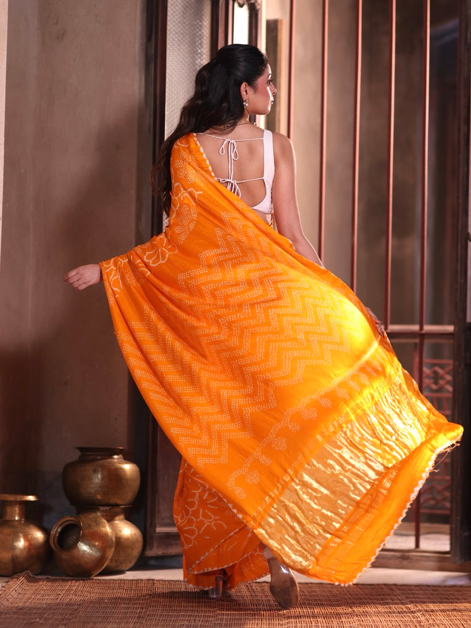 Woman in an orange saree with a white blouse standing in a room with decorative pots.