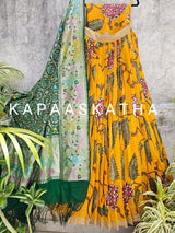 Yellow floral dress with green dupatta on a textured wall background, featuring the brand 'Kapaaskatha'.
