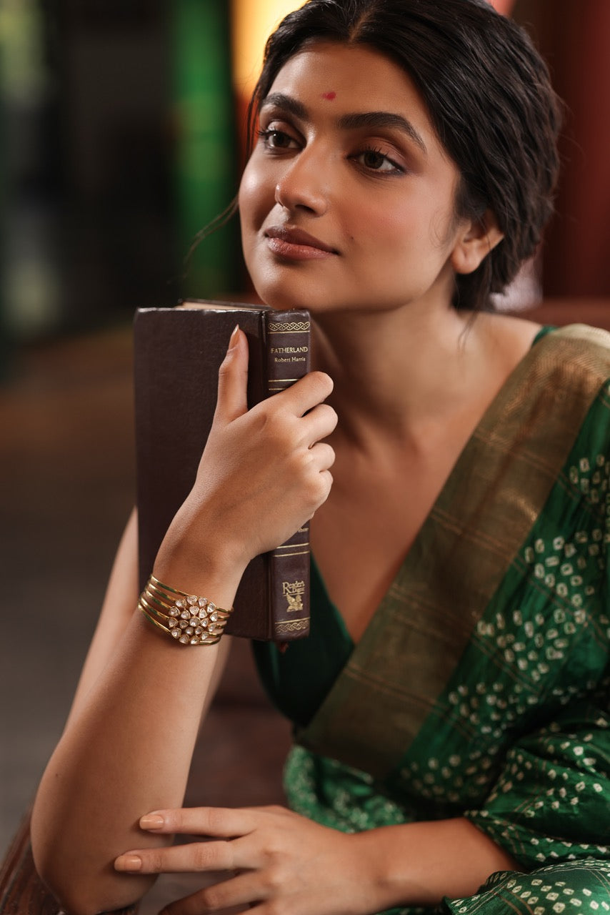 Woman in a green saree holding a cup, sitting on a wooden bench wearing a 92.5 silver handcuff with moissanite polki.