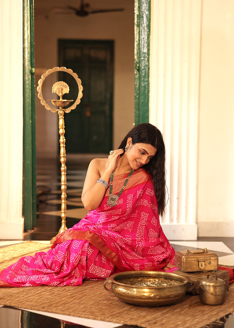 Woman in a pink saree sitting on a mat with brass items around her, in front of a mirror.